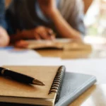 Un cahier et un stylo sur une table d'étude, avec des apprentis travaillant ensemble en arrière-plan flou, symbolisant le programme d'aide apprenti 2024