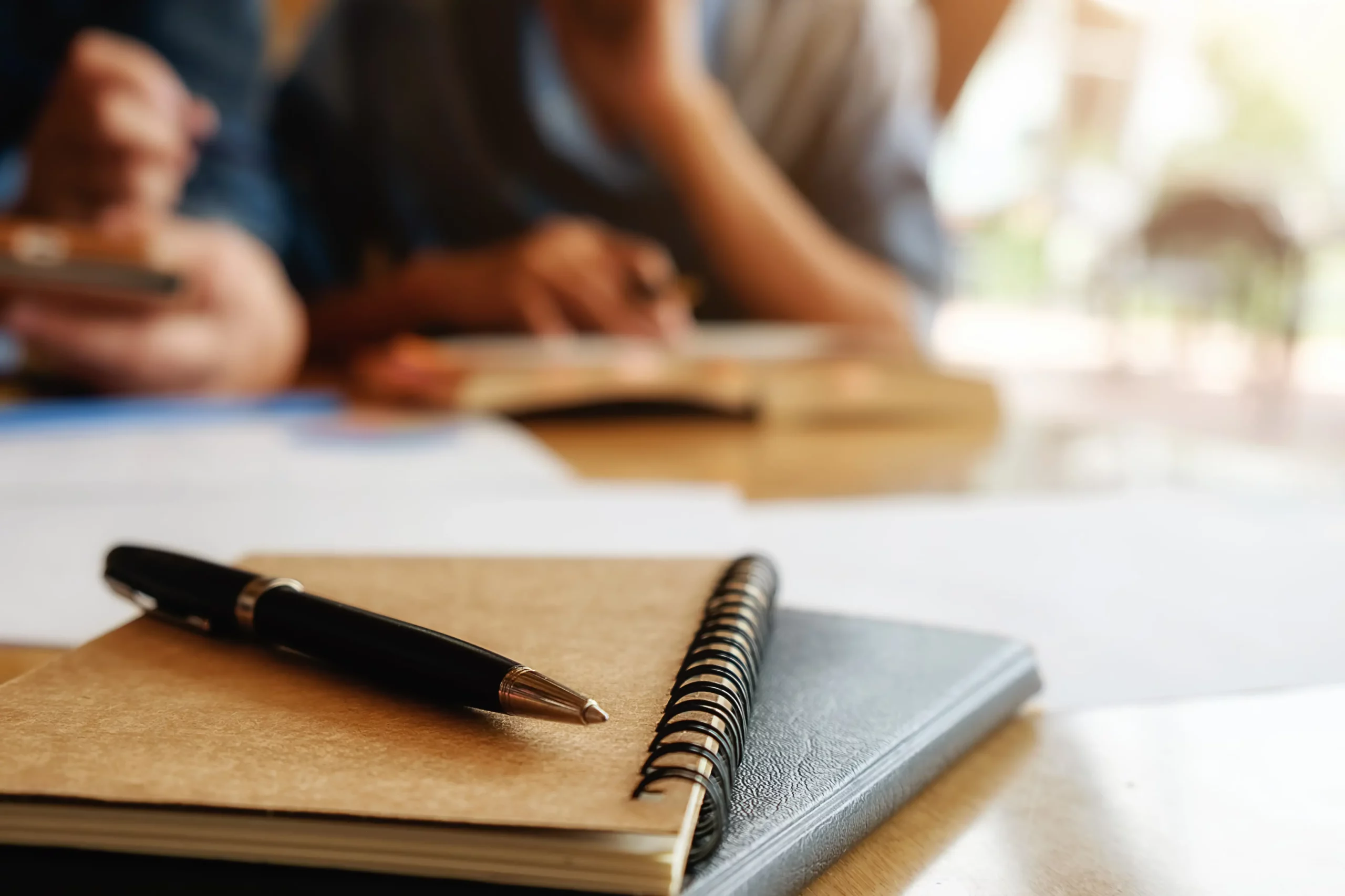 Un cahier et un stylo sur une table d'étude, avec des apprentis travaillant ensemble en arrière-plan flou, symbolisant le programme d'aide apprenti 2024